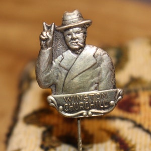 May include: Silver-toned lapel pin featuring a portrait of Winston Churchill in a hat, giving a peace sign. The pin has the text "Winston Churchill" on a banner below the portrait.