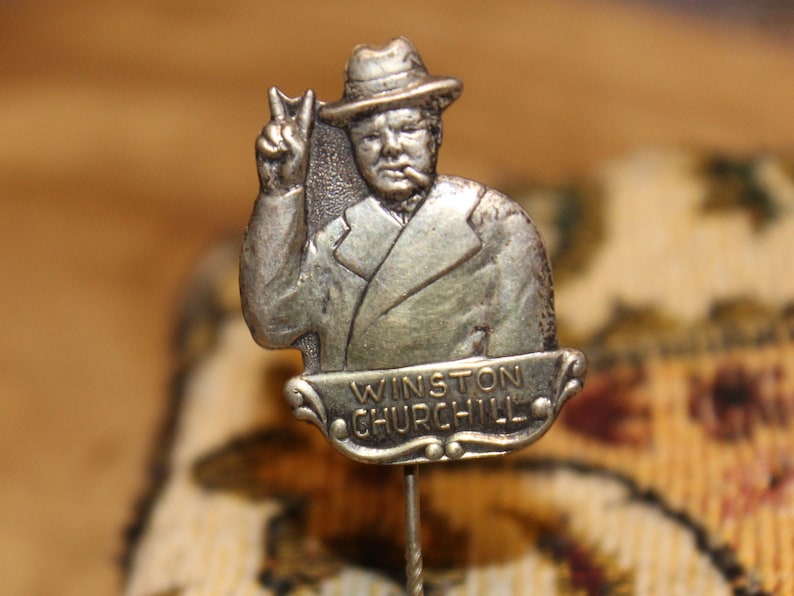 May include: Silver-toned lapel pin featuring a portrait of Winston Churchill in a hat, giving a peace sign. The pin has the text "Winston Churchill" on a banner below the portrait.