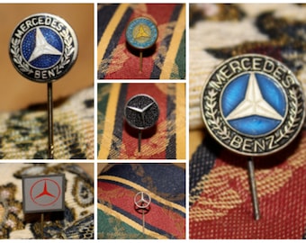 May include: Five different Mercedes-Benz pins with varying designs. The pins are silver, black, and blue with white accents. The pins feature the Mercedes-Benz logo, a three-pointed star.