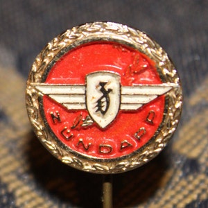 May include: A gold-colored pin with a red and white enamel design. The design features a shield with a stylized letter "J" inside, flanked by white wings. The words "N.U.V.A.D" are written around the shield in a circular pattern.
