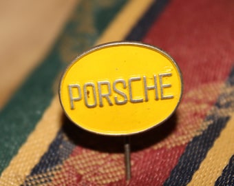 May include: A yellow oval pin with the word "PORSCHE" in silver lettering. The pin has a silver border and a silver pin backing. The background is a colorful plaid pattern.