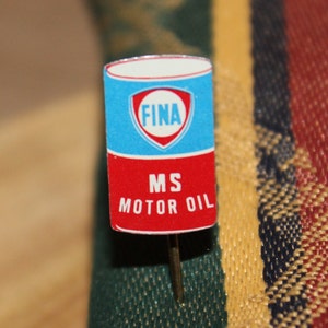 May include: A vintage pin featuring a rectangular design with a blue top section displaying the word "FINA" inside a white and red emblem. The bottom section is red with the text "MS MOTOR OIL". The pin has a silver needle.
