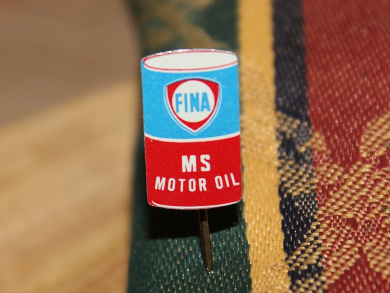 May include: A vintage pin featuring a rectangular design with a blue top section displaying the word "FINA" inside a white and red emblem. The bottom section is red with the text "MS MOTOR OIL". The pin has a silver needle.