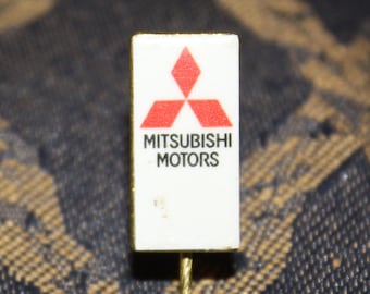 May include: A white pin with a red Mitsubishi Motors logo. The logo is a three-pointed red star.