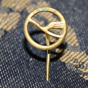 May include: A gold-colored pin with a stylized bird design inside a circle. The pin has a long, thin stem.