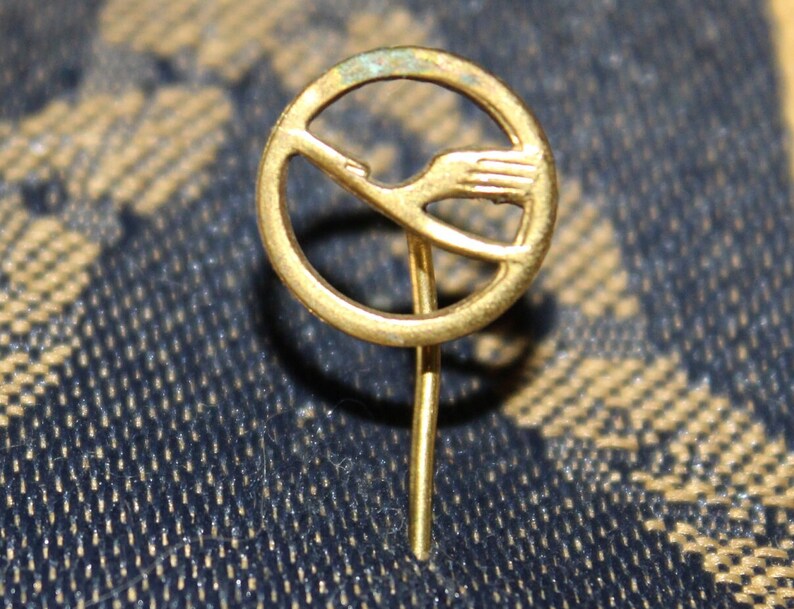 May include: A gold-colored pin with a stylized bird design inside a circle. The pin has a long, thin stem.