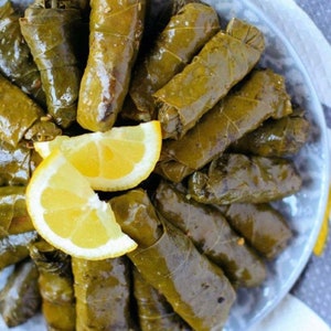 May include: A close-up of a plate of grape leaves stuffed with rice and herbs, also known as dolmades. The grape leaves are a vibrant green color and are arranged in a circular pattern. There are two slices of lemon on top of the grape leaves.