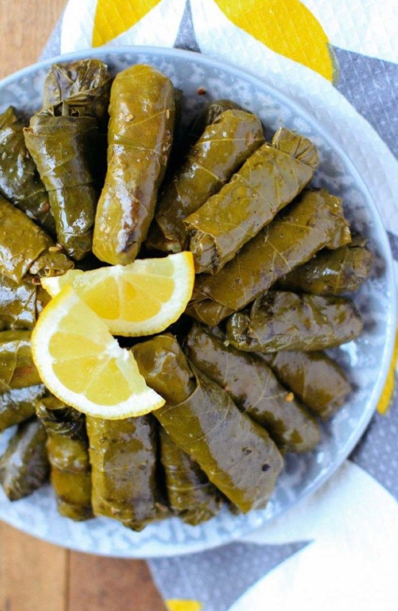 May include: A close-up of a plate of grape leaves stuffed with rice and herbs, also known as dolmades. The grape leaves are a vibrant green color and are arranged in a circular pattern. There are two slices of lemon on top of the grape leaves.
