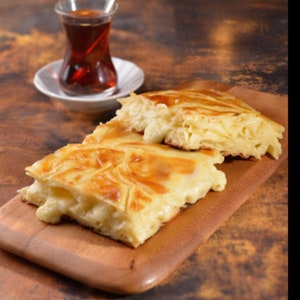 May include: Two pieces of golden brown pastry with a white cheese filling on a wooden cutting board. A glass of tea is in the background.