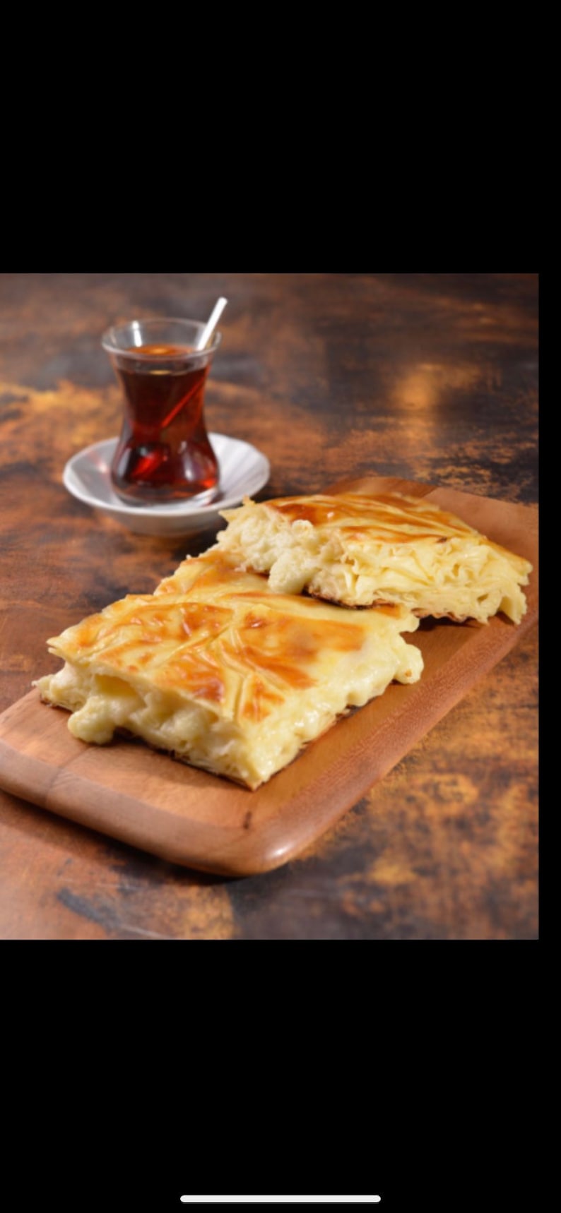 May include: Two pieces of golden brown pastry with a white cheese filling on a wooden cutting board. A glass of tea is in the background.