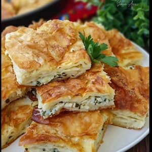 May include: A plate of golden brown, flaky pastry squares filled with cheese and herbs. The pastry is arranged in a stack, with some pieces overlapping. There is a sprig of parsley on top of the pastry.