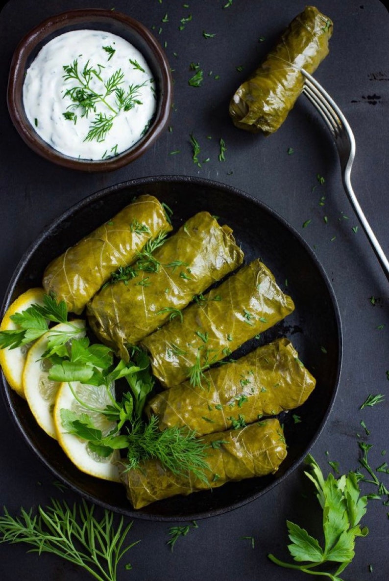 May include: A black skillet filled with grape leaves stuffed with rice and herbs. The grape leaves are arranged in a circular pattern. There is a small bowl of white sauce and a fork on the side.