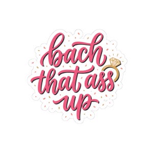 May include: A die-cut sticker featuring the pink cursive text "bach that ass up" with a gold diamond ring icon replacing the letter "o" in the word "bach". The design is surrounded by small pink and gold confetti dots on a white background.