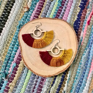 Puede incluir: Un par de pendientes de madera con un dise&ntilde;o circular y una franja de hilo rojo y dorado.