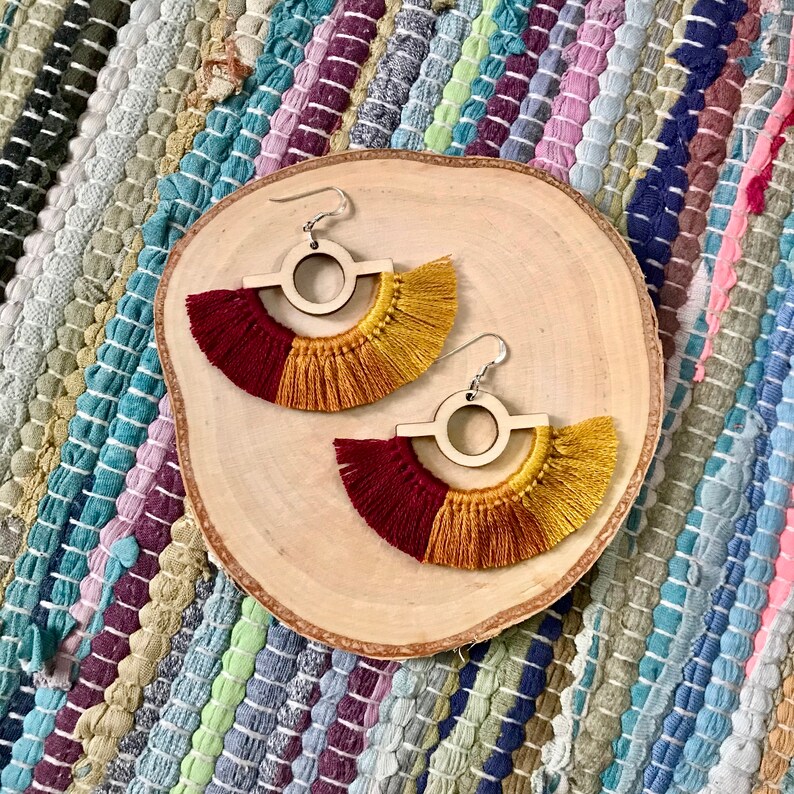 Puede incluir: Un par de pendientes de madera con un dise&ntilde;o circular y una franja de hilo rojo y dorado.