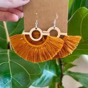 Puede incluir: Un par de pendientes de borla de color amarillo mostaza con un c&iacute;rculo de madera y una barra recta que conecta los dos c&iacute;rculos. Los pendientes tienen ganchos de plata.