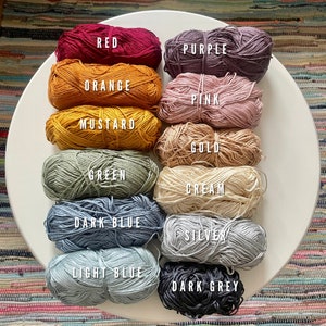 Puede incluir: Una colecci&oacute;n de ovillos de lana en varios colores, incluyendo rojo, naranja, mostaza, verde, azul oscuro, azul claro, gris oscuro, morado, rosa, dorado, crema y plateado. Los ovillos de lana est&aacute;n dispuestos en un c&iacute;rculo sobre una superficie blanca.