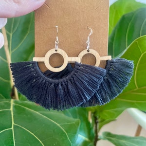 Puede incluir: Un par de pendientes de borla gris oscuro con formas geom&eacute;tricas de madera. Los pendientes presentan un c&iacute;rculo con un c&iacute;rculo m&aacute;s peque&ntilde;o cortado en el centro y una barra horizontal que se extiende desde el c&iacute;rculo.