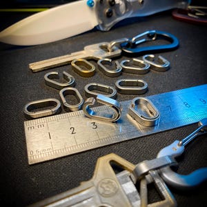 May include: A collection of small, oval-shaped metal split rings and connectors scattered on a metal ruler. The ruler shows measurements in millimeters and centimeters. Two keys are attached to carabiners, and a folding knife is visible in the background.