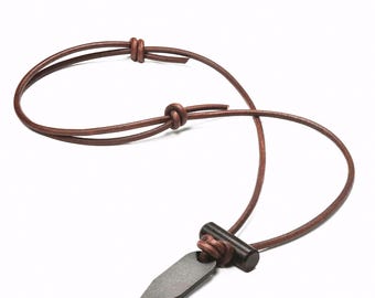 May include: A necklace featuring a dark gray, flat, geometric metal pendant attached to a dark brown leather cord. The cord includes a small, cylindrical dark wood bead and is secured with adjustable sliding knots for a customizable length.