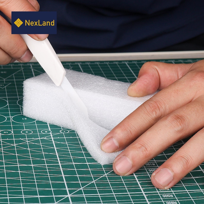 May include: A white craft knife cutting a white foam block on a green cutting mat. The knife is held by a person's hands. The logo "NexLand" is in the upper left corner.
