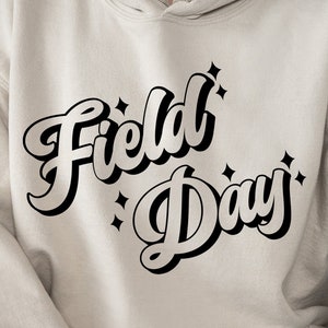 May include: A beige hoodie with the text "Field Day" in a black retro font. The text is stylized with a sparkle design.
