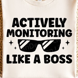 May include: A white t-shirt with black text that reads "Actively Monitoring Like A Boss" with a pair of sunglasses and two stars on either side of the sunglasses.