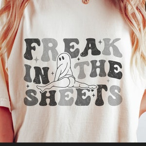 May include: A white t-shirt with a retro-style graphic design featuring a ghost in a bed with the text "Freak in the Sheets".