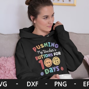 Pode incluir: Um moletom preto com o texto "PUSHING My teacher's BUTTONS FOR 100 Days" em cores do arco-&iacute;ris com bot&otilde;es coloridos ao redor do texto.
