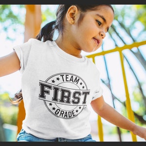 May include: A white t-shirt with a black and white graphic that says "Team First Grade" in a circle.