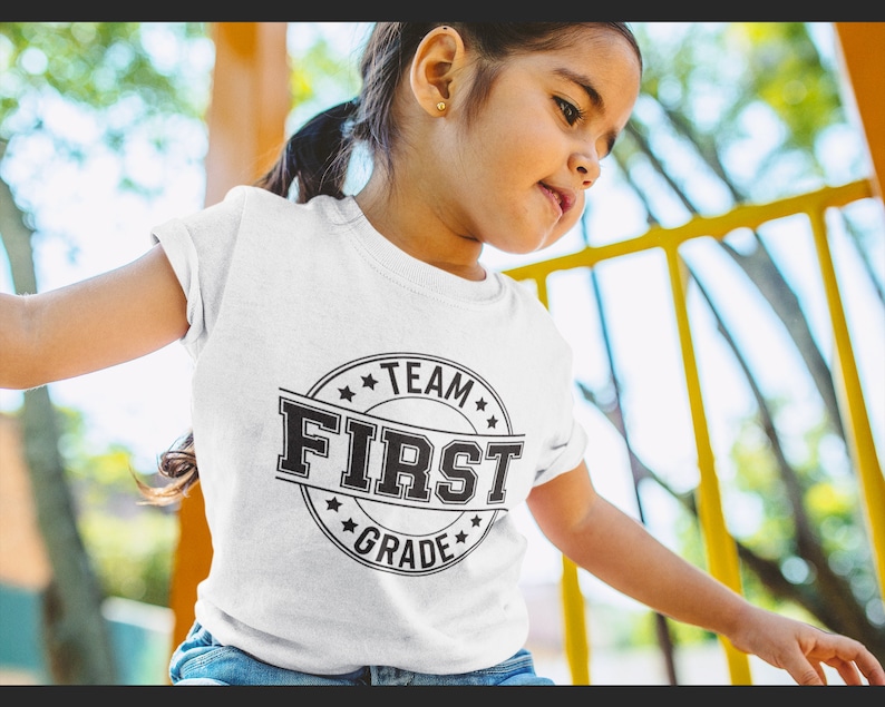 May include: A white t-shirt with a black and white graphic that says "Team First Grade" in a circle.