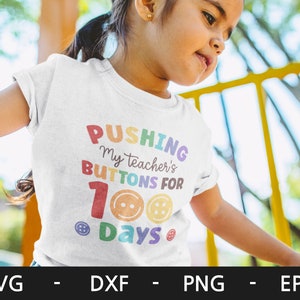 Pode incluir: Uma camiseta branca com o texto "Pushing my teacher's buttons for 100 days" em cores do arco-&iacute;ris com designs de bot&otilde;es.