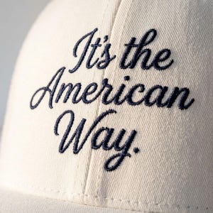 Puede incluir: Gorra de béisbol color crema con la frase bordada en azul marino "It's the American Way". La gorra tiene un diseño clásico con visera curva, adecuada para el uso diario.