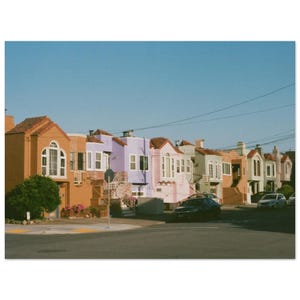 May include: A row of colorful houses with orange, purple, and pink facades under a clear blue sky. Each house has a different design, with red tile roofs and white window frames. Cars are parked on the street in front of the houses.