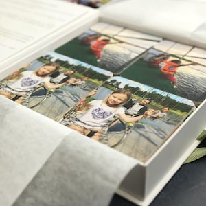 Peut inclure: Une boîte blanche avec un ruban vert, contenant un ensemble de photographies imprimées. Les photos présentent une jeune fille sur une balançoire, un groupe de personnes sur un bateau et un groupe de personnes dans un parc.