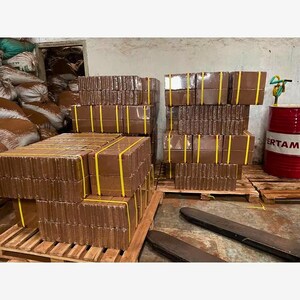 Puede incluir: Varias pilas de bloques rectangulares marrones, asegurados con correas amarillas, est&aacute;n dispuestas sobre palets de madera. Los bloques est&aacute;n cuidadosamente organizados, lo que sugiere un producto listo para la distribuci&oacute;n. Un barril rojo con texto blanco es visible en el fondo.