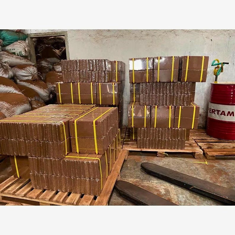 Puede incluir: Varias pilas de bloques rectangulares marrones, asegurados con correas amarillas, est&aacute;n dispuestas sobre palets de madera. Los bloques est&aacute;n cuidadosamente organizados, lo que sugiere un producto listo para la distribuci&oacute;n. Un barril rojo con texto blanco es visible en el fondo.