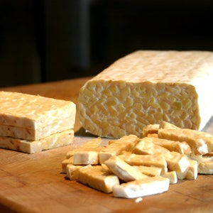 May include: A close-up of tempeh, a fermented soybean product, cut into different sizes. The tempeh is light brown and has a slightly grainy texture. It is arranged on a wooden chopping board.