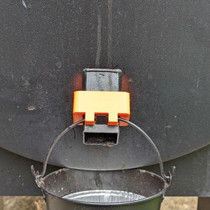May include: A black metal smoker with a yellow plastic drip pan attached to the bottom. A black bucket with a handle is positioned underneath the drip pan to catch the drippings.