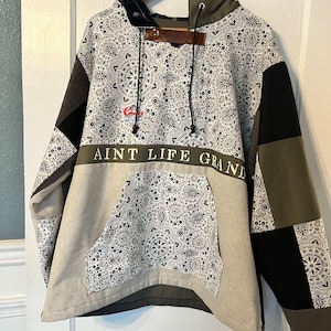 May include: A patchwork hoodie with a black and white patterned design, a green hood, and the text "AINT LIFE GRAND" in white on a green band.