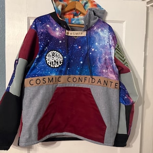 May include: A patchwork hoodie with a galaxy print on the hood and chest. The hoodie features a large front pocket and the words "COSMIC CONFIDANTE" across the chest. Patches of red, gray, and black fabric are sewn together.