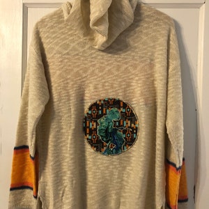 Peut inclure: Un pull en maille beige à col roulé et manches longues. Le pull a un grand patch rond sur le devant avec un motif géométrique coloré. Les manches ont des rayures orange et bleues.