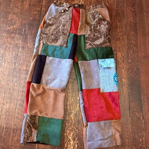 May include: Patchwork pants with a variety of fabrics in shades of brown, green, red, and gray. The pants have a drawstring waist and a loose fit. The design features a mix of textures and patterns, creating a unique and colorful look.