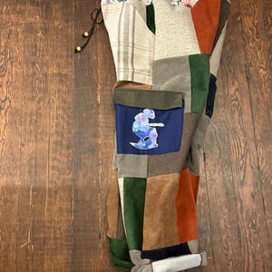 May include: Patchwork pants in a variety of textures and colors, including corduroy, suede, and patterned fabrics. The pants feature a blue pocket with a graphic design. The waistband has a drawstring closure.