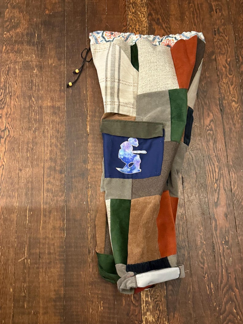 May include: Patchwork pants in a variety of textures and colors, including corduroy, suede, and patterned fabrics. The pants feature a blue pocket with a graphic design. The waistband has a drawstring closure.