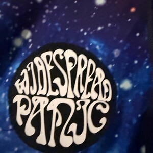 Puede incluir: Primer plano de una tela con un patr&oacute;n de galaxia en tonos azules, morados y blancos. Un parche circular negro con las palabras "WIDESPREAD PANIC" en letras blancas estilizadas est&aacute; centrado en la tela.