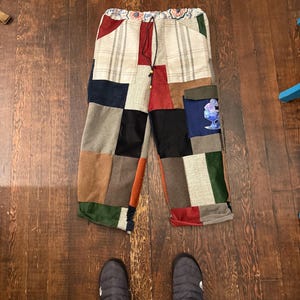 May include: Patchwork pants with a drawstring waist. The pants feature a variety of fabric patches in colors like beige, red, green, and black. The pants are laying on a wooden floor. Gray slippers are at the bottom of the image.