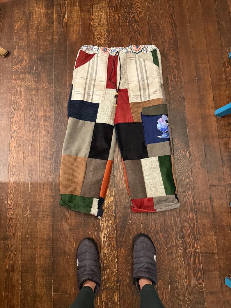 May include: Patchwork pants with a drawstring waist. The pants feature a variety of fabric patches in colors like beige, red, green, and black. The pants are laying on a wooden floor. Gray slippers are at the bottom of the image.
