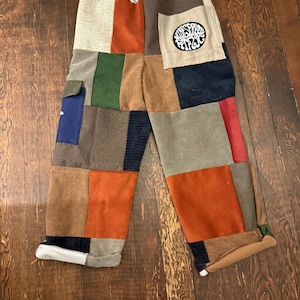 May include: Patchwork pants with a variety of corduroy and suede patches in shades of brown, orange, green, and blue. The pants have a drawstring waist and a pocket with the text "Wildwood Circus".