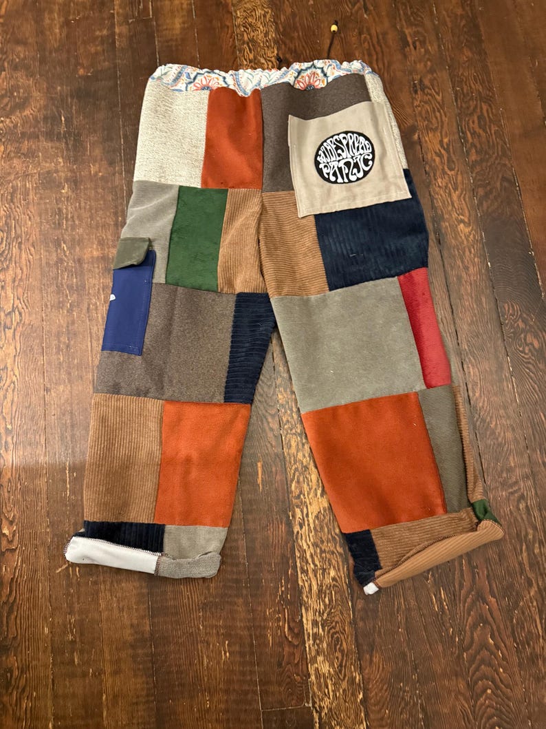 May include: Patchwork pants with a variety of corduroy and suede patches in shades of brown, orange, green, and blue. The pants have a drawstring waist and a pocket with the text "Wildwood Circus".